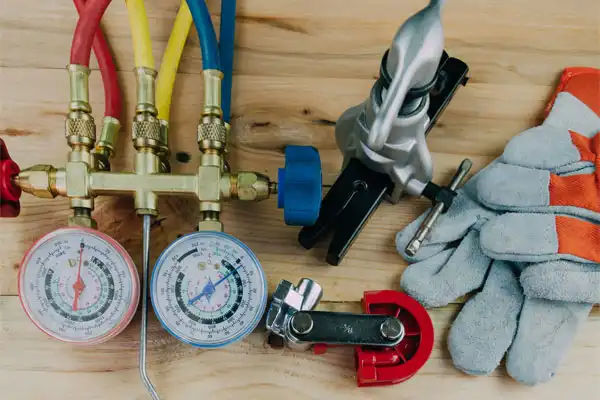 HVAC tools on a wooden surface, including a manifold gauge set with red, blue, and yellow hoses, pipe cutter, vice, and protective gloves.
