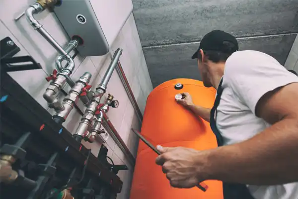 A worker in a cap adjusts a valve on a large orange tank in a utility room with exposed pipes.