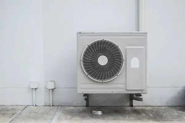 A wall-mounted air conditioning unit with a visible fan and metal casing against a light gray wall.