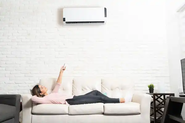 Person lying on a couch, pointing a remote control at an air conditioner mounted on a white brick wall.